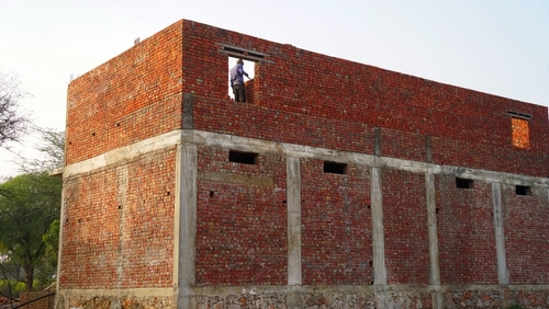A artisan standing in the window of an under-construction site brick building, working to shape the structure. Person at work exterior bricklayer wall with cement. Builder, engineer, worker