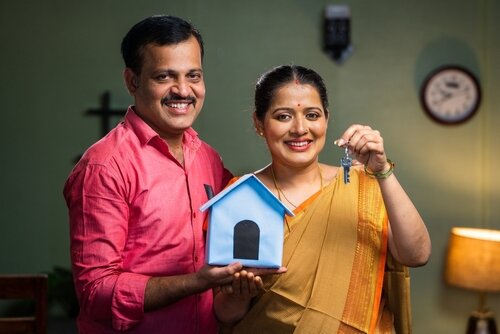 Happy smiling couple showing new house keys by toy home while looking camera - concept of new home purchasing, home loan and investment.