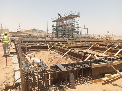 Construction site with workers assembling steel reinforcement, wooden formwork, and scaffolding for a concrete foundation