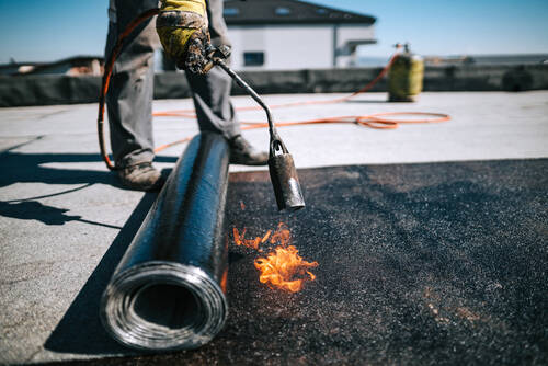 Professional workers insulating rooftop with bitumen membrane. Waterproofing details at construction site. Bituminous membrane waterproofing system details and installation on flat rooftop.