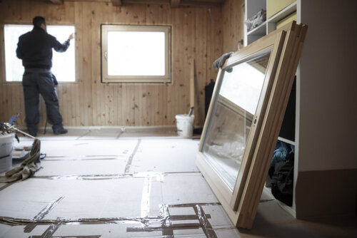 Worker in the background installing new, three pane wooden windows in an old wooden house, with a new window in the foreground. Home renovation, sustainable living, energy efficiency concept. 