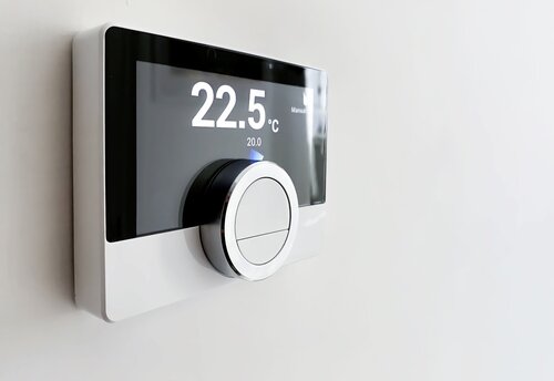 Screen of a smart digital thermostat with the external and target temperatures indicated. Rotary dial to control and adjust. Scheduling and programmable domestic heating. White wall and black frame